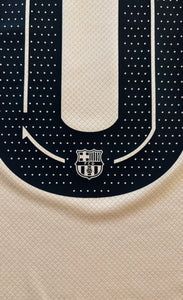 Close-up of a sports jersey with a logo on the chest against a dark blue and beige background. Yamal Kobe Barcelona
